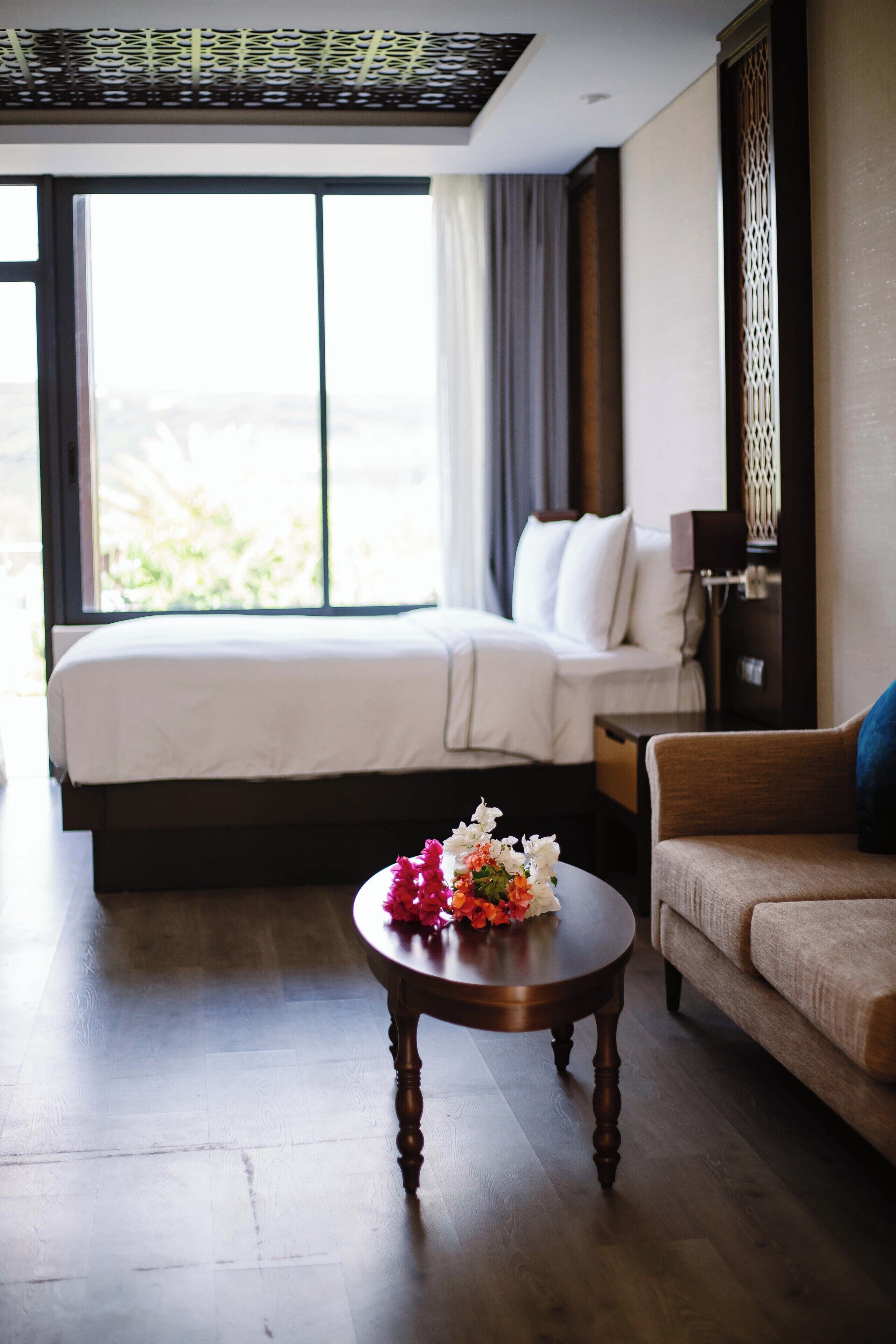 Quarto de hotel com uma cama, um sofá, uma mesa redonda de madeira com flores e uma janela grande com cortinas.