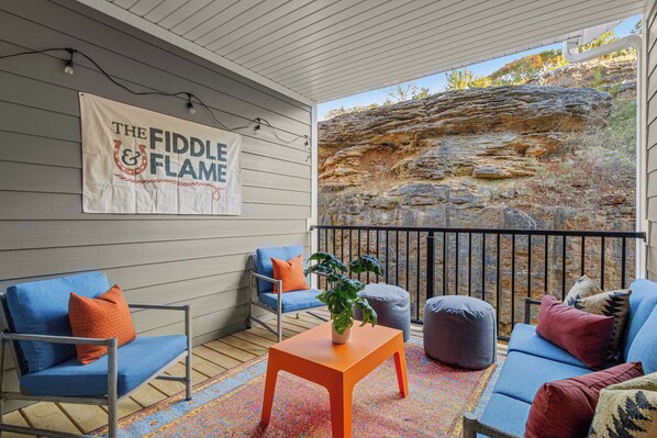 Un espacio de estar al aire libre cubierto con mobiliario azul y gris, una alfombra colorida y un cartel con la inscripción 'THE FIDDLE FLAME'.
