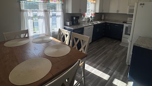 Uma cozinha com armários brancos, uma mesa de jantar de madeira com jogos americanos e uma janela com cortinas florais.