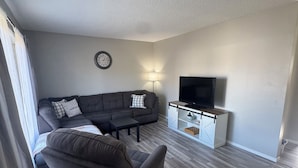 Uma sala de estar com sofá seccional, uma poltrona, televisão e um pequeno armário.