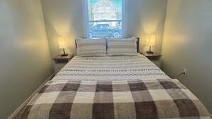 Quarto com colcha xadrez, duas luminárias de cabeceira, janela com persianas e uma tomada visível na parede.