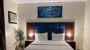 A hotel room with a large bed, two bedside tables, a nightstand with a lamp, a wall-mounted air conditioner, a potted plant, and a painting of a snowy landscape. - fakhamat almasif (Taif)