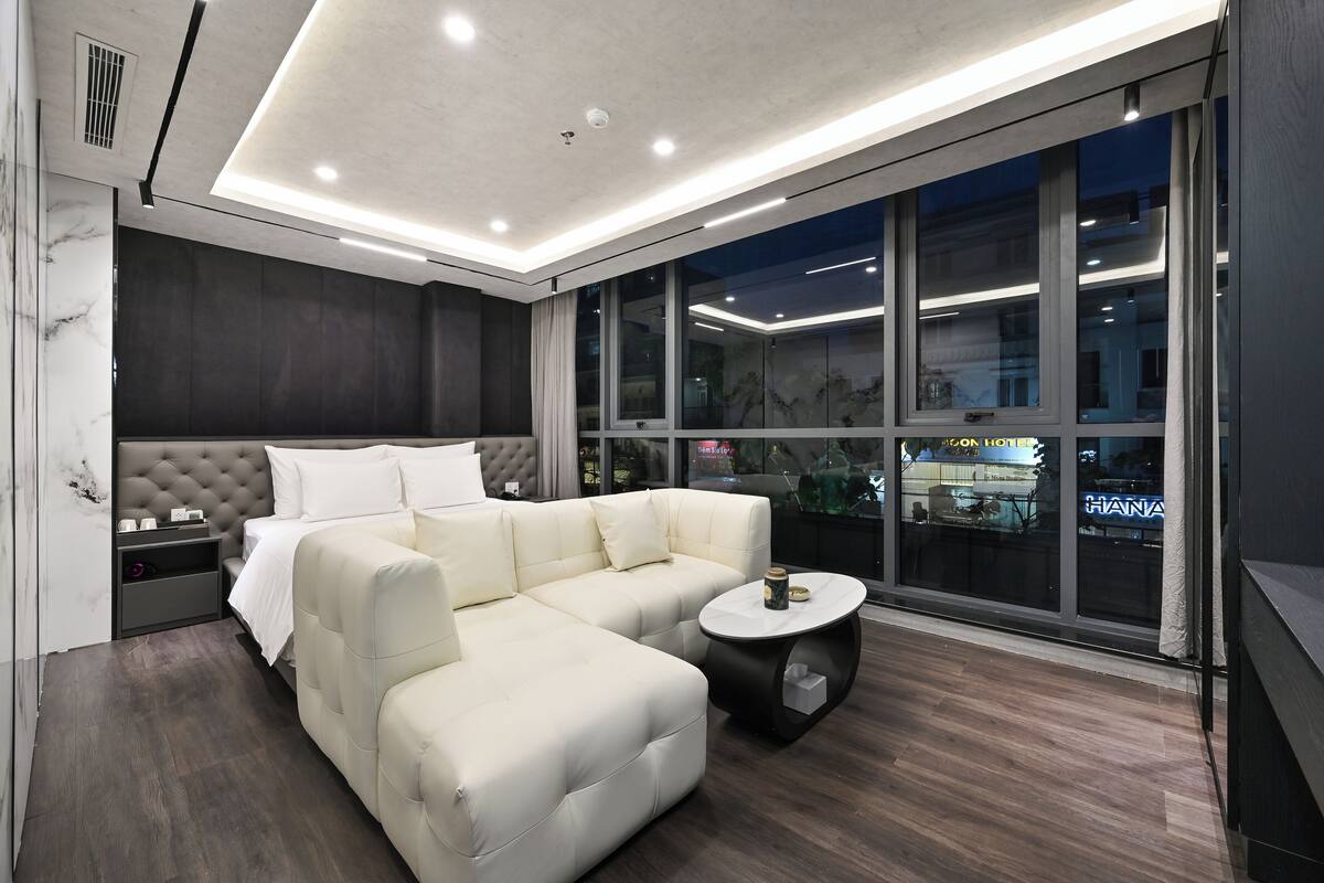 Une chambre d’hôtel moderne avec un canapé d’angle, une table basse ronde et une grande fenêtre donnant sur la ville.