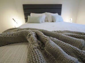 Un letto con una coperta a maglia e cuscini in una stanza con una parete spoglia e una testiera scura.