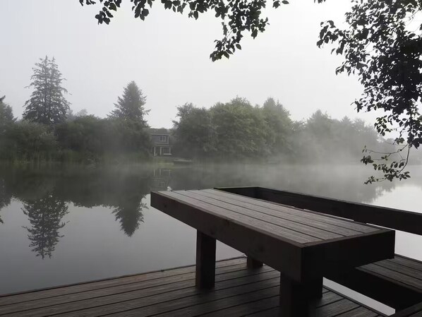 Un molo in legno con una panchina che si affaccia su un lago calmo, circondato da alberi e un edificio in lontananza.