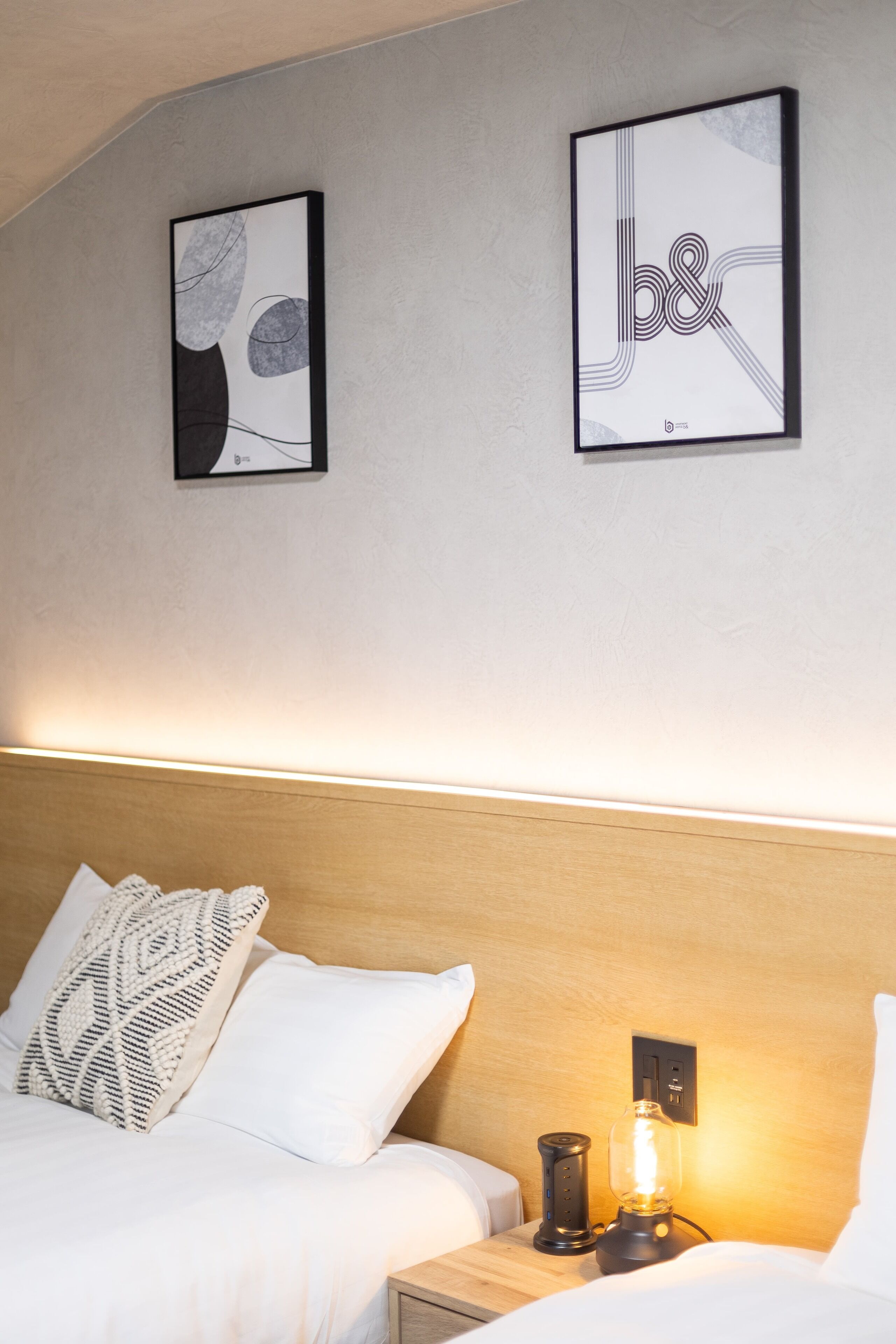 Ein Hotelzimmer mit einem Bett, zwei Kissen, einem Nachttisch mit Lampe sowie zwei gerahmten Kunstwerken an der Wand.