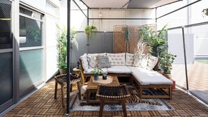 Un espace extérieur aménagé, abrité et doté d’un canapé confortable, de tables et de chaises en bois, entouré de plantes en pot et offrant une vue sur la rue.