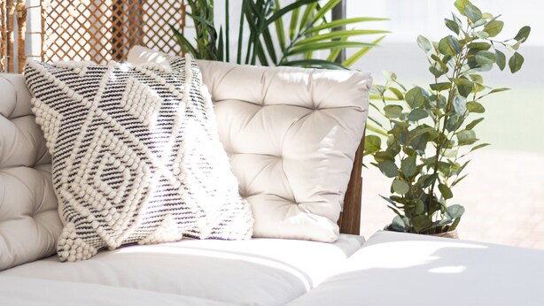 Un canapé d’angle avec un coussin blanc et un coussin décoratif à motifs, une petite plante en pot et un paravent en osier.