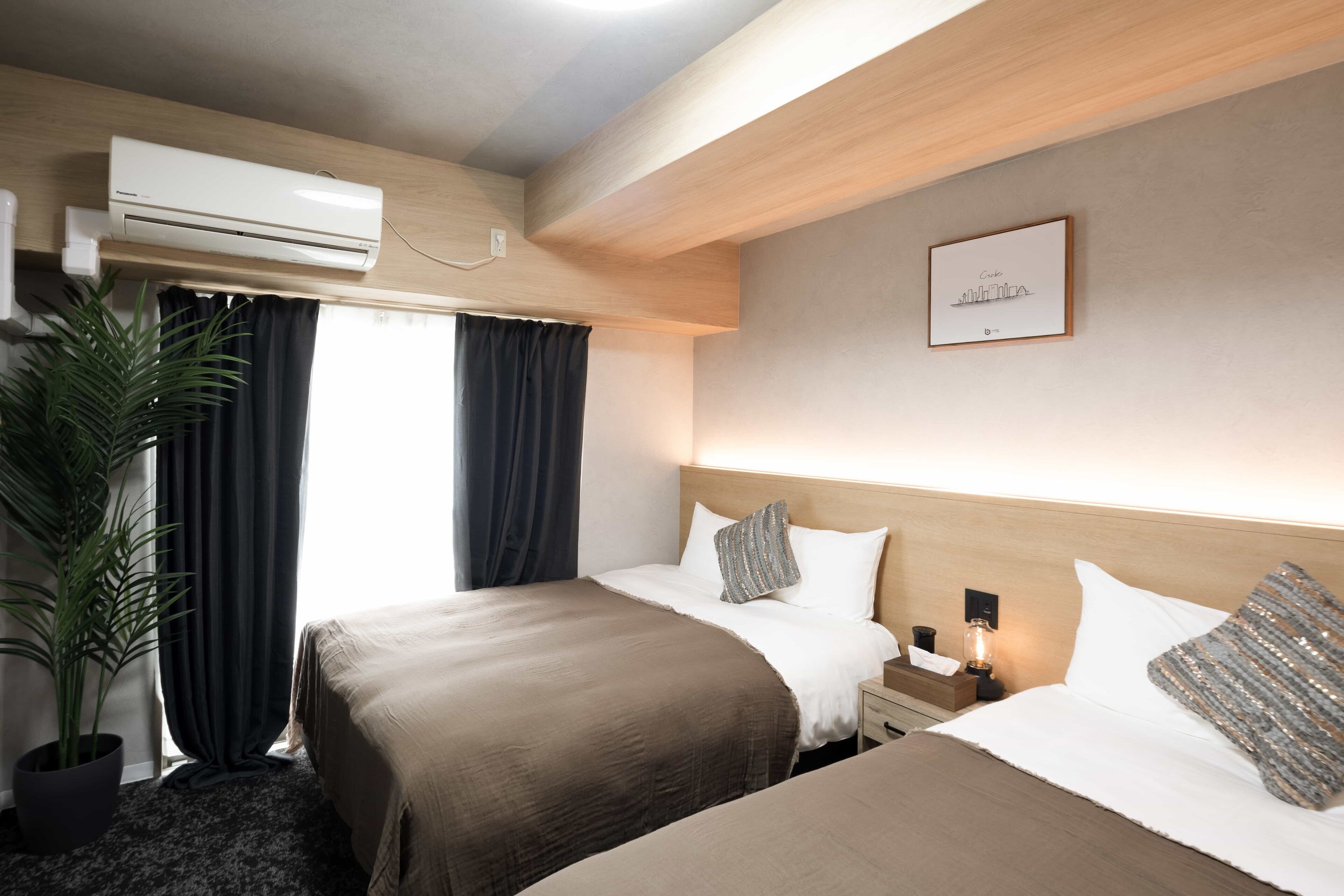 Ein Hotelzimmer mit zwei Betten, einem Blumentopf, einer Klimaanlage, einem Nachttisch mit Lampe und einem gerahmten Bild an der Wand.