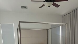 Une chambre moderne avec un lit à baldaquin, une table de chevet, une commode, un ventilateur de plafond et une fenêtre avec des stores.