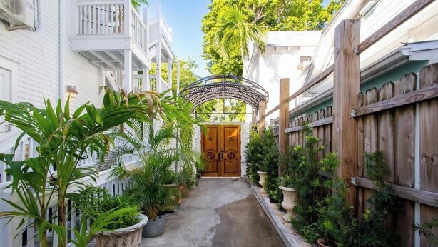 Un camí estret que condueix a una porta de fusta amb un arc decoratiu, envoltat de plantes en testos i una tanca de fusta.