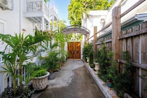 A narrow pathway leading to a wooden door with a decorative archway, surrounded by potted plants and a wooden fence. - Cabana on Duval  |  Monthly studio w/ shared pool (Key West)