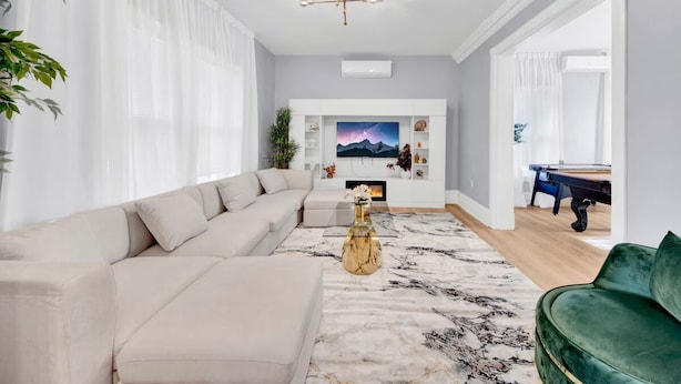 A modern living room with a beige sectional sofa, a green armchair, a marble-patterned rug, a gold coffee table, and a flat-screen TV mounted on a white wall.