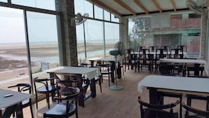 A restaurant with large windows overlooking a beach, wooden tables and chairs, and a fan. - Sampan Resort (Cox's Bazar)