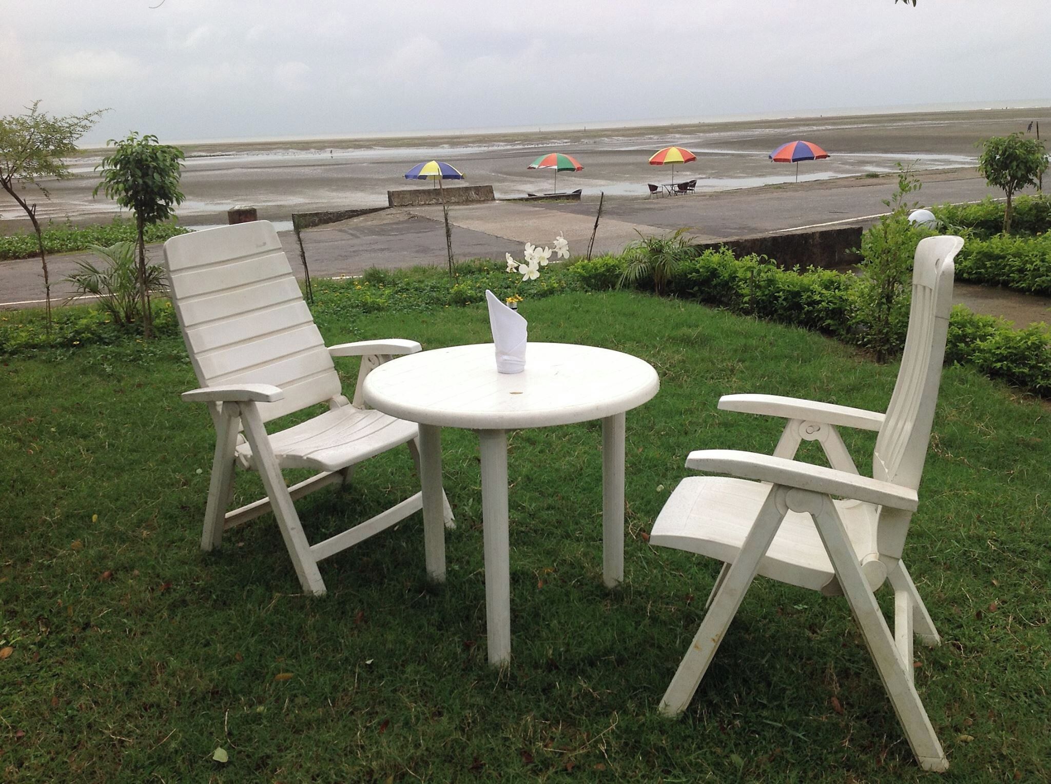 A grassy outdoor seating area with two white chairs and a small round table, overlooking a beach with umbrellas.