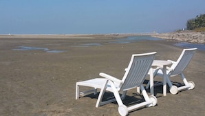 Two white beach chairs on a sandy beach with a calm sea and a rocky shoreline in the background. - Sampan Resort (Cox's Bazar)