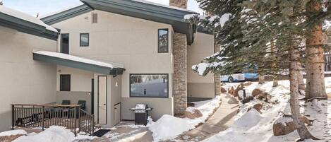 A modern building with a stone chimney, a covered patio with outdoor seating, and a snow-covered landscape.