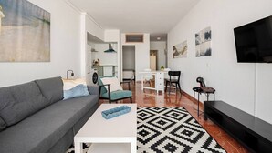 A modern living room with a grey sofa, a white coffee table, and a patterned rug. There is a dining area with a white table and chairs, and a kitchen in the background. - Plaza de la Magdalena II (Seville)