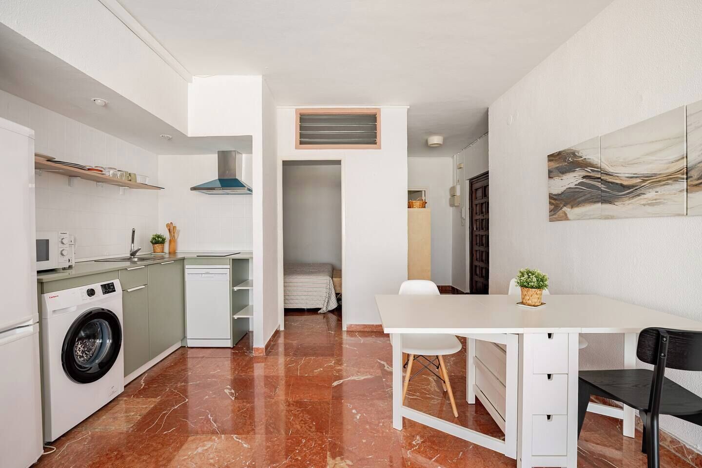 Una cocina compacta con lavadora, un comedor con mesa y sillas, y una pequeña sala de estar con cama.
