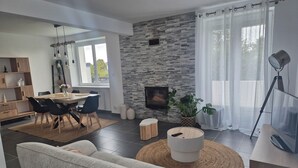 Ett modernt vardagsrum med en vägg av sten, en öppen spis, en matplats med ett träbord och stolar samt ett stort fönster med tunna, genomskinliga gardiner.