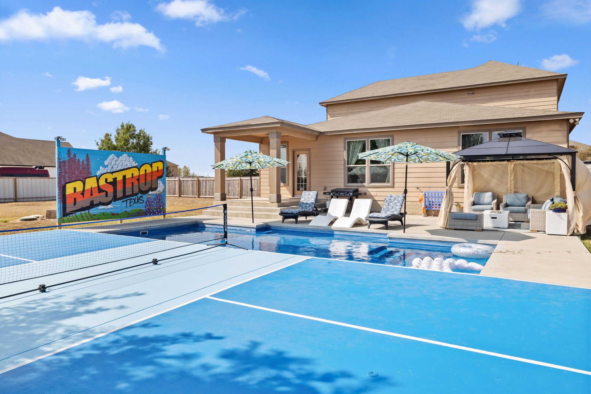 A residential swimming pool with lounge chairs and umbrellas, a covered seating area, and a house in the background.