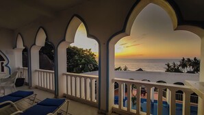 Varanda com vista para o pôr do sol sobre o mar, com piscina e espreguiçadeiras.