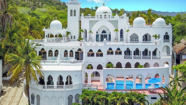 Um edifício grande, branco e de vários andares, com janelas em arco e telhados em cúpula, rodeado por vegetação exuberante e um céu azul límpido.