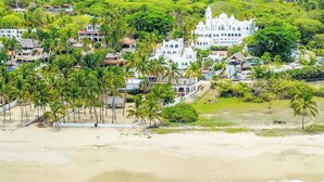 Uma área costeira com praia, palmeiras e um edifício branco grande com várias janelas e uma torre.
