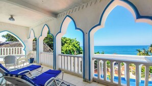 Uma área externa coberta com mobiliário azul e branco, chão de azulejos e vista para o mar.
