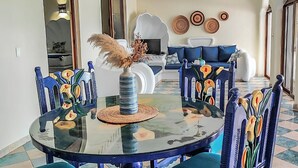 Uma área de refeições com mesa de vidro cercada por cadeiras coloridas, um vaso com flores secas e um sofá com almofadas estampadas ao fundo.