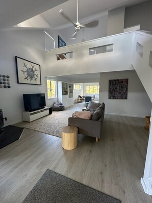 A spacious living area with a flat-screen TV, a grey sofa, a wooden coffee table, and a staircase leading to an upper level.