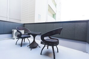 A balcony with two wicker chairs and a small table, an air conditioning unit, and a white building in the background. - b&FLAT-Hanazono (Osaka)