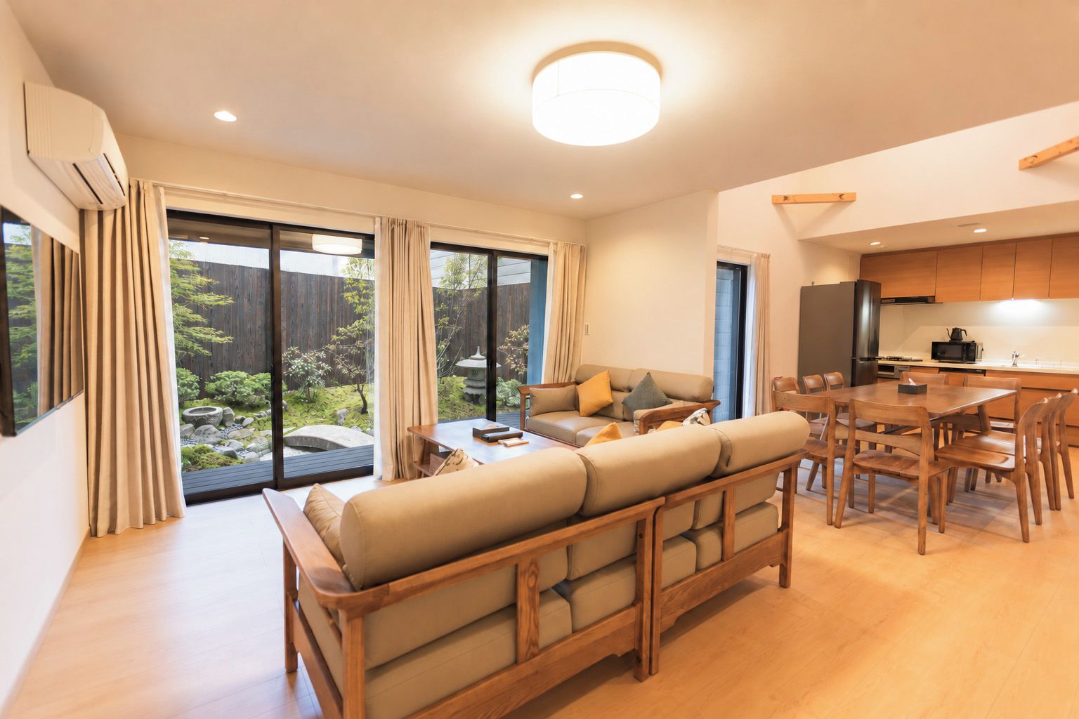 Una sala de estar moderna con sofá, mesa de comedor y sillas. Hay una zona de cocina con electrodomésticos y vistas a un jardín a través de ventanas amplias.