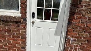 A white door with a warning sign for security cameras, a window with a view of trees, and a brick wall.