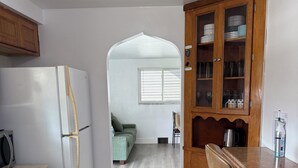 A kitchen with a refrigerator, wooden cabinets, a dining table with chairs, and a view into a living room with a sofa and a window.