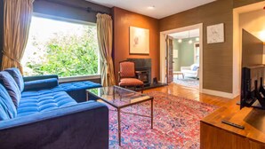 A modern living room with a blue sofa, a glass coffee table, and a patterned rug. There is a fireplace, a television, and a door leading to another room.