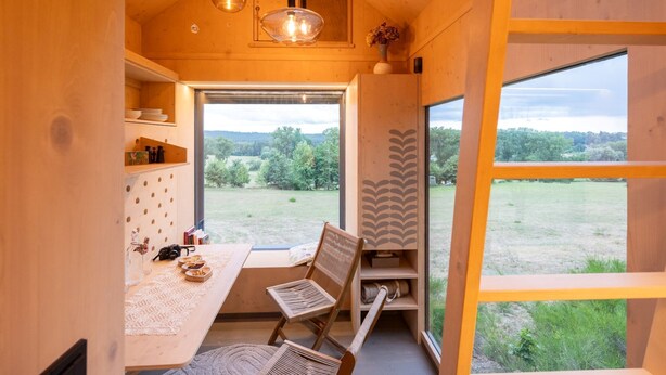 Un interior de madera acogedor con comedor, estantería y un amplio ventanal con vistas a un paisaje verde.