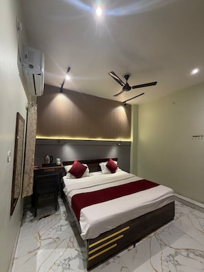 A hotel room with a bed, a desk, a chair, a ceiling fan, and an air conditioning unit. - Jogi Lodge (Varanasi)