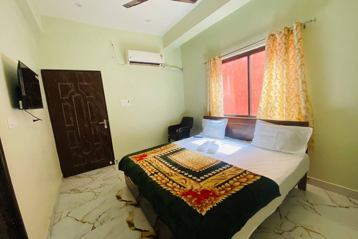 A hotel room with a bed, a ceiling fan, an air conditioning unit, a window with curtains, and a door.