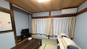 Een kamer met een houten plafond, een televisie op een standaard, een bank met kussens, een tafel en een raam met gordijnen.