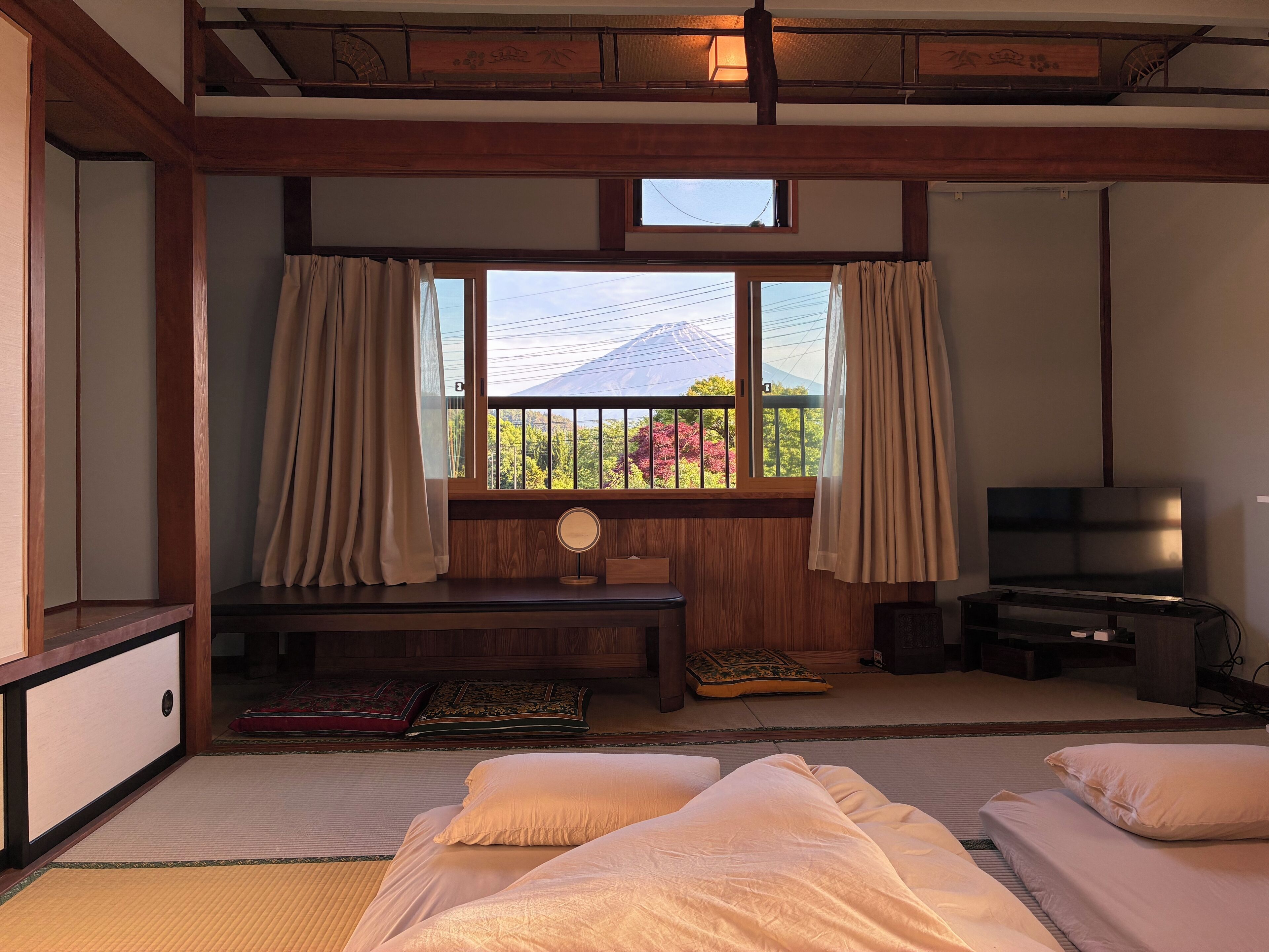 Una habitación de estilo japonés tradicional con suelo de tatami, una cama baja de madera, un televisor y un ventanal con cortinas.
