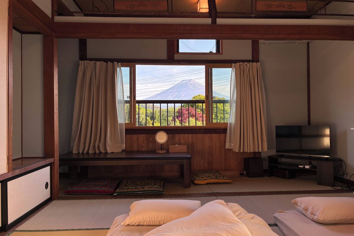 Ett traditionellt japanskt rum med tatamimattor på golvet, en låg säng av trä, en TV och ett fönster med gardiner.