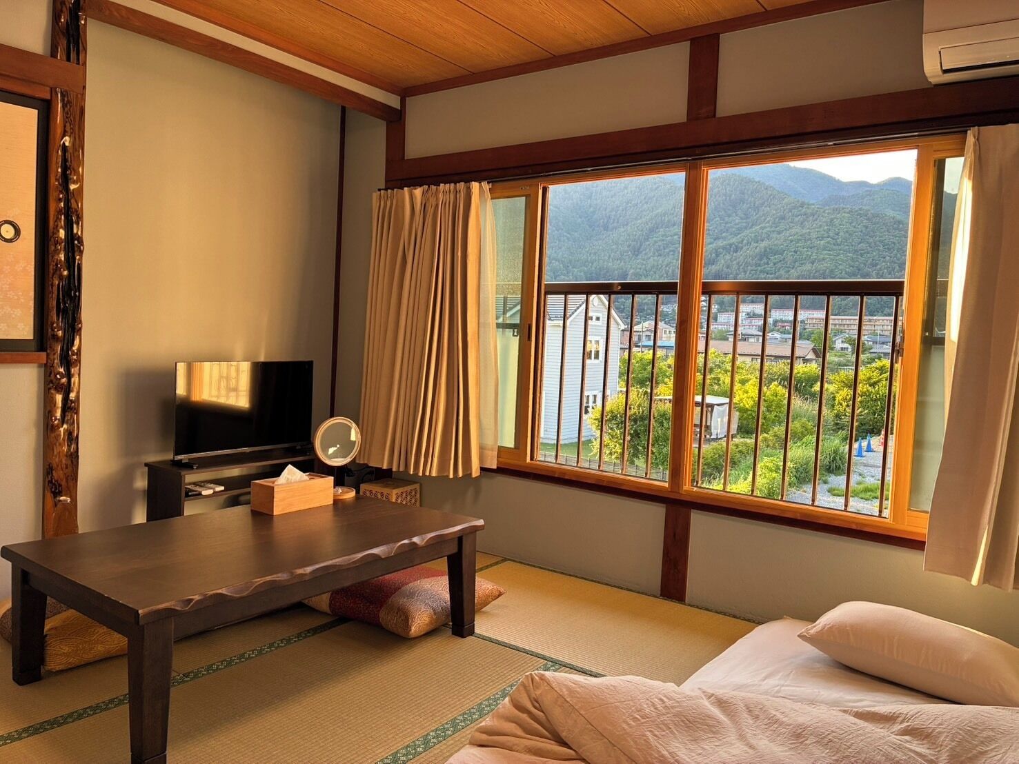 Una habitación con suelo de tatami, televisión, mesita de noche, espejo y un gran ventanal con vistas a un paisaje montañoso.
