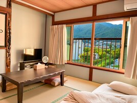 A room with a bed, a TV, a table, a mirror, and a window with a view of a mountain and a lake.