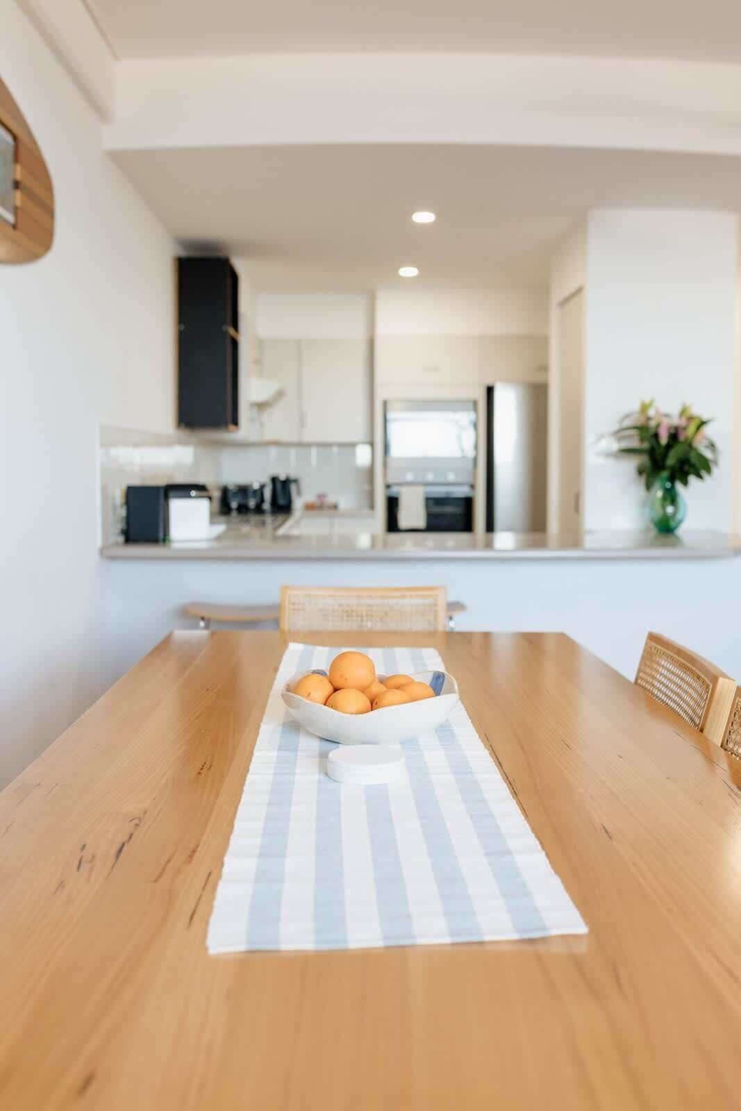 A dining table with a bowl of oranges on a striped placemat, a wooden chair, and a vase with flowers on a table in a modern kitchen setting.