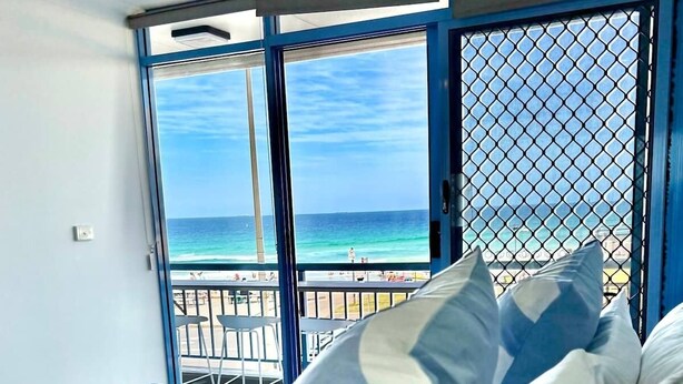 A hotel room with a bed, pillows, a ceiling fan, and a view of the beach through a window. - Beachfront Apartment | Pool, Tennis & Parking (Newcastle East)