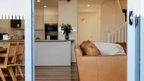 A modern living room with a sofa, a dining area with wooden chairs, a kitchen in the background, and a ceiling fan. - Beachfront Apartment | Pool, Tennis & Parking (Newcastle East)