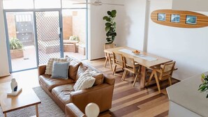 A modern living room with a brown leather sofa, a wooden dining table with chairs, and a large window with a view of the outside. - Beachfront Apartment | Pool, Tennis & Parking (Newcastle East)