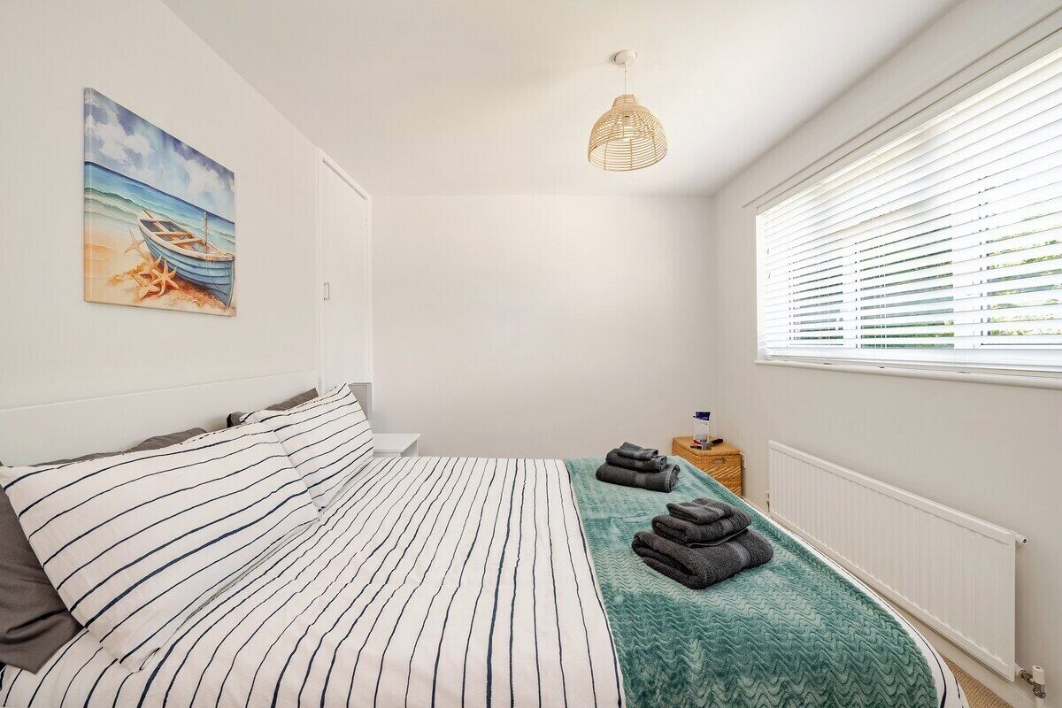 Une chambre avec un lit à rayures, un tableau représentant une scène de plage, un luminaire en osier et une fenêtre avec des stores.