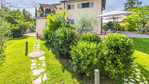 Una casa a due piani con un vialetto fiorito che conduce all'ingresso, circondata da vegetazione lussureggiante e un patio con mobili da esterno.